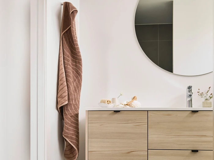 Et brunt håndklæde hænger på en krog ved siden af et badeværelsesbord i lyst træ med et rundt spejl over og små pyntegenstande på bordet.