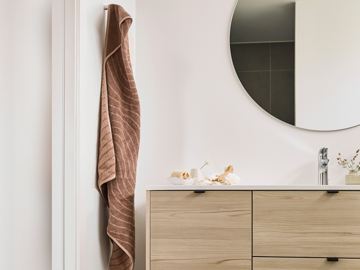 Et brunt håndklæde hænger på en knage ved siden af et rundt spejl over et badeværelsesbord i lyst træ med en vask og små pyntegenstande.