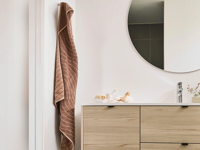 Et brunt håndklæde hænger på en knage ved siden af et rundt spejl over et badeværelsesbord i lyst træ med en vask og små pyntegenstande.