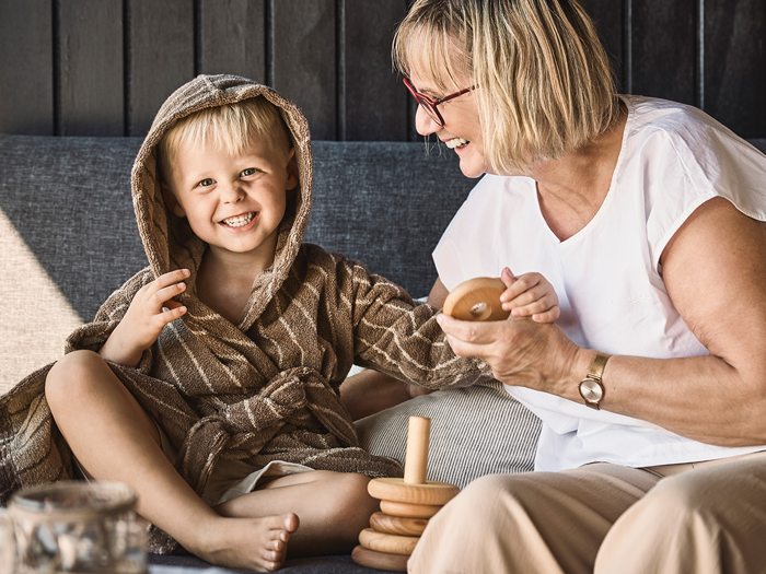 Et smilende barn i badekåbe sidder ved siden af en ældre kvinde med briller, og begge leger med trælegetøj, der kan stables, i en sofa.