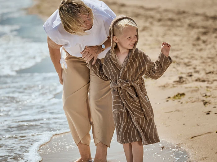 En voksen og et barn i en kappe med hætte går barfodet langs stranden og smiler, mens de nyder vandet og sandet.