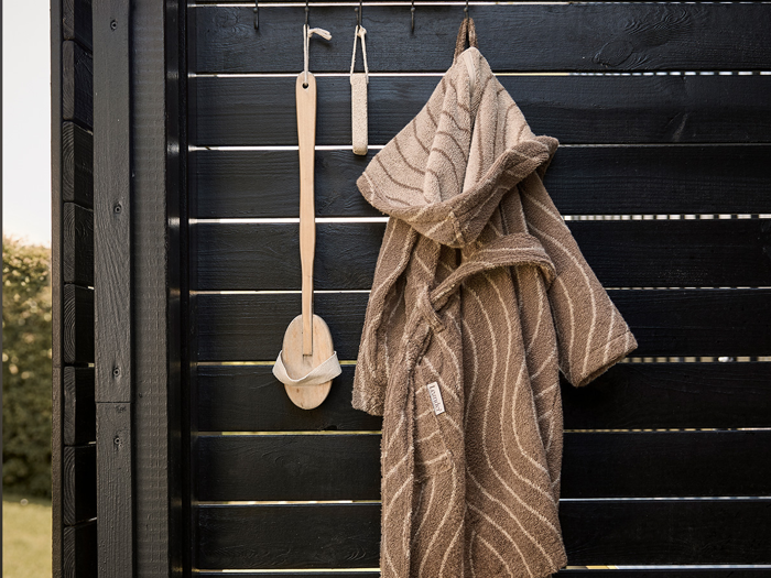 En brun badekåbe og badebørster af træ hænger på kroge mod en sort lamelvæg af træ.