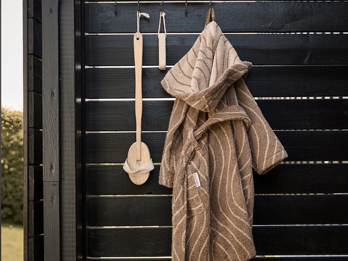 En brun badekåbe og badebørster af træ hænger på kroge mod en sort lamelvæg af træ.