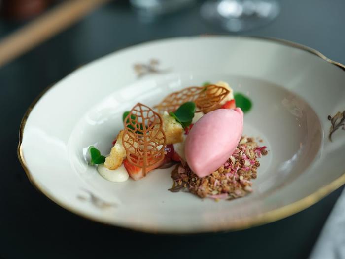 En anrettet dessert med pink sorbet, crumble, jordbær, mikrogrønt, fløde og dekorative karamelliserede sukkerstykker på et hvidt fad med guldkant.