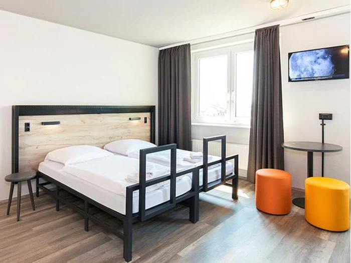 Moderne hotelværelse med dobbeltseng, lyst trægulv, et lille rundt bord, to orange taburetter, to sideborde og et væghængt tv ved et vindue med grå gardiner.