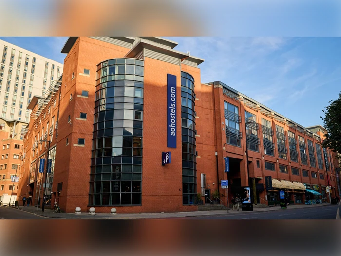 En moderne rød murstensbygning med store glasvinduer og et blåt aohostels.com-skilt på hjørnet, beliggende på en bygade under en klar himmel.