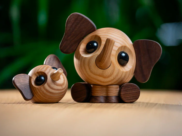 To søde elefantfigurer af træ med runde kroppe, store sorte øjne og mørke ører sidder på en træoverflade med en sløret grøn baggrund.