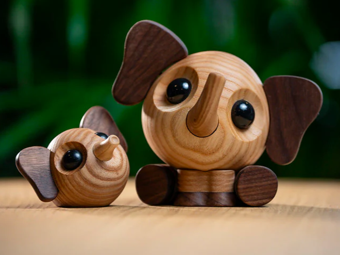 To runde elefantfigurer af træ, en siddende og en liggende, med store mørke ører og skinnende sorte øjne, på en træplade med sløret grøn baggrund.