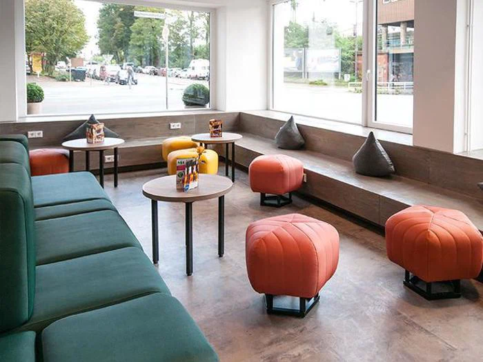 Moderne lounge med grønne polstrede bænke, orange ottomaner, runde borde, grå puder og store vinduer, der giver naturligt lys og udsigt til gaden udenfor.