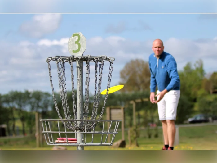 En mand i blå jakke kaster en gul disc mod en discgolfkurv markeret med tallet 3 på en græsklædt udendørs bane.