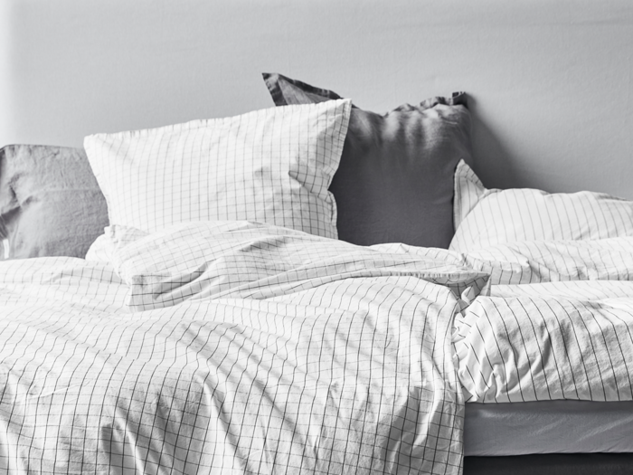 En pænt redt seng med hvidt og gråt gittermønstret sengetøj og puder mod en lysegrå væg.