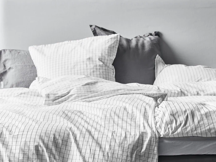 En pænt redt seng med hvidt og gråt gittermønstret sengetøj og puder mod en lysegrå væg.