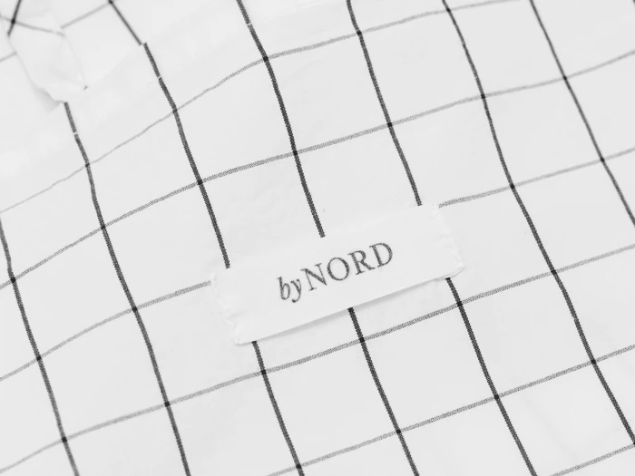 Nærbillede af et hvidt stof med et sort gittermønster og en påsyet etiket med byNORD i sort tekst.