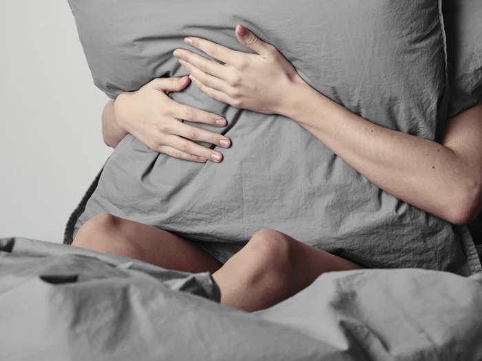 En person sidder på en seng og holder om en stor grå pude, der delvist dækker kroppen. Kun arme, hænder og ben er synlige, og der er gråt sengetøj i forgrunden.