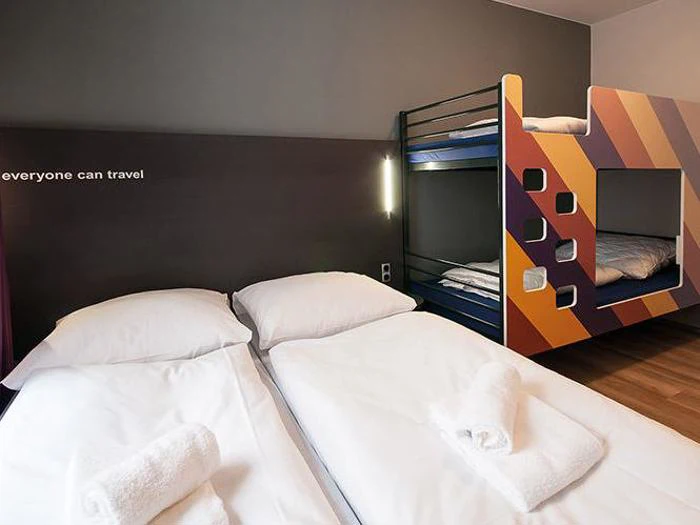 Et moderne hostelværelse med to enkeltsenge, en farverig køjeseng, håndklæder på hver seng og ordene everyone can travel på væggen.