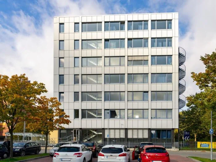 En moderne kontorbygning i seks etager med store vinduer, parkerede biler foran, træer på begge sider og en udvendig vindeltrappe til højre.