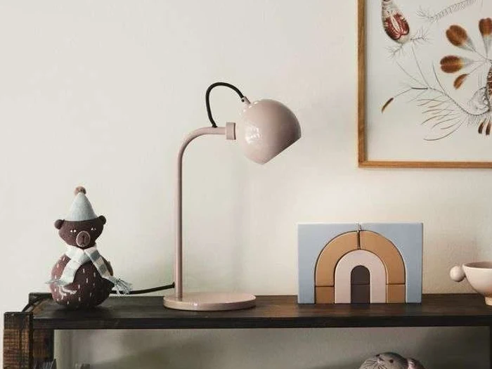 En lyserød skrivebordslampe står på en hylde ved siden af en brun bamse med blå hat, en legetøjsregnbue og et indrammet kunstværk med tørrede planter på en hvid væg.