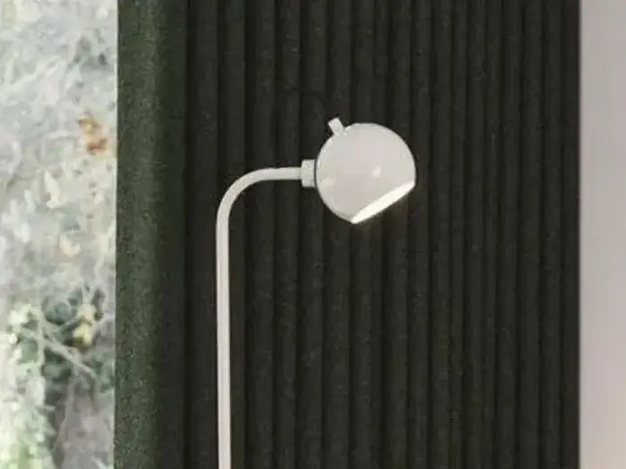 En moderne hvid gulvlampe med et rundt hoved står foran en mørk, lodret struktureret væg ved siden af et vindue med udsigt til træer udenfor.
