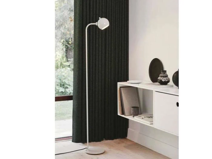 En moderne hvid gulvlampe står nær et vindue med mørke gardiner ved siden af et hvidt skab med bøger, tallerkener og en dekorativ vase.