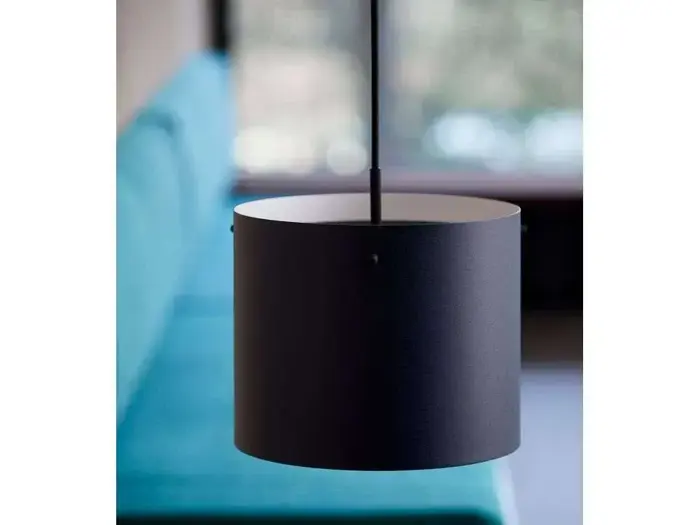 En sort cylindrisk pendel hænger foran en sløret blå sofa og vinduesbaggrund.