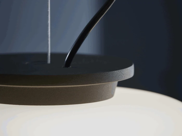 Nærbillede af en moderne pendellampe med matsort top, ophængningsledninger og en lysende hvid skærm mod en mørk baggrund.