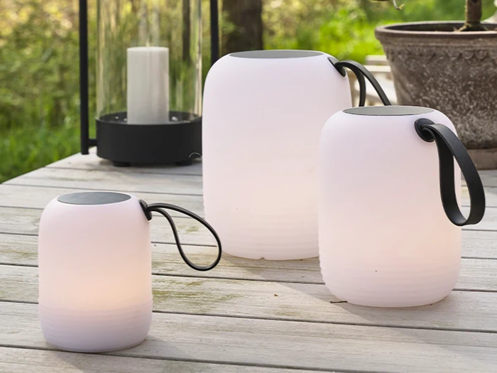 Tre hvide lanterner i forskellige størrelser med sorte håndtag er placeret på en træflade udendørs i nærheden af en urtepotte og en stor lyslygte.