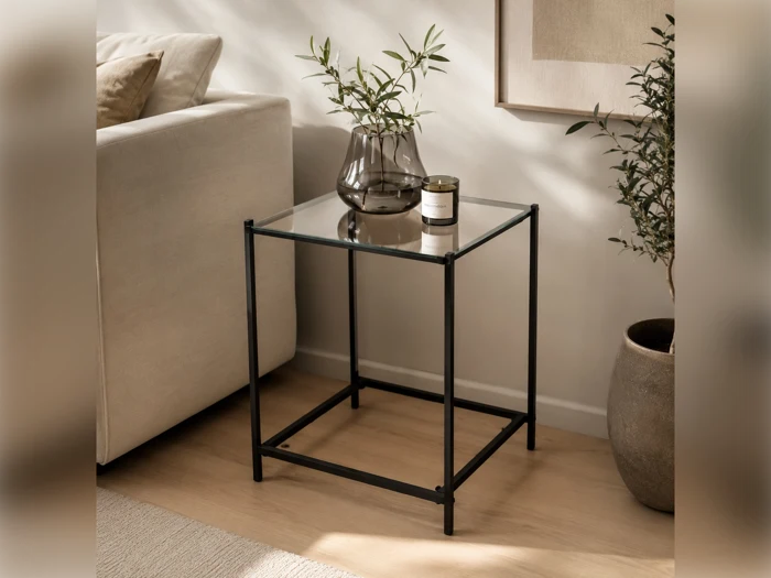 Et minimalistisk sidebord i glas med en sort metalramme, hvorpå der står en vase med grønne planter og et stearinlys, ved siden af en beige sofa og en stor potteplante på et trægulv.