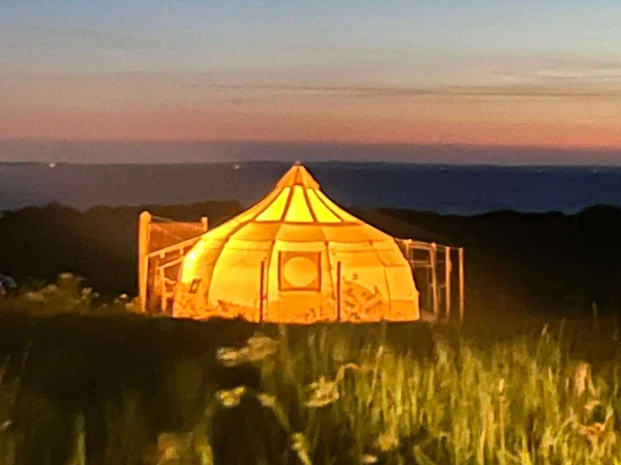 En lysende gul jurte er opstillet på en græsmark i skumringen, med en farverig solnedgang og mørke bakker i baggrunden.