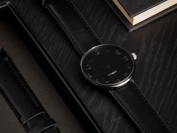 Et elegant sort armbåndsur med minimalistisk design og læderrem hviler på en mørk træoverflade ved siden af en bog.