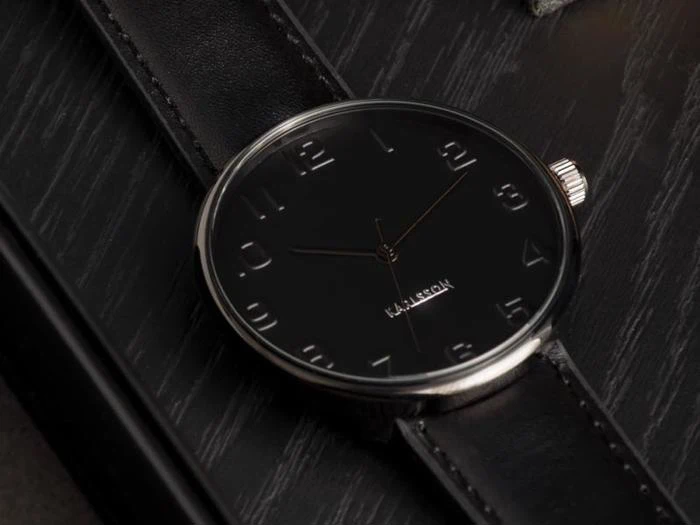 Et nærbillede af et elegant sort armbåndsur med sølvfarvede tal og visere, placeret på en sort læderrem over en mørk træoverflade.