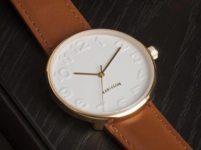 Et guldindfattet armbåndsur med hvid urskive, ophøjede tal, brun læderrem og navnet Karlsson på urskiven, der hviler på en mørk træplade.