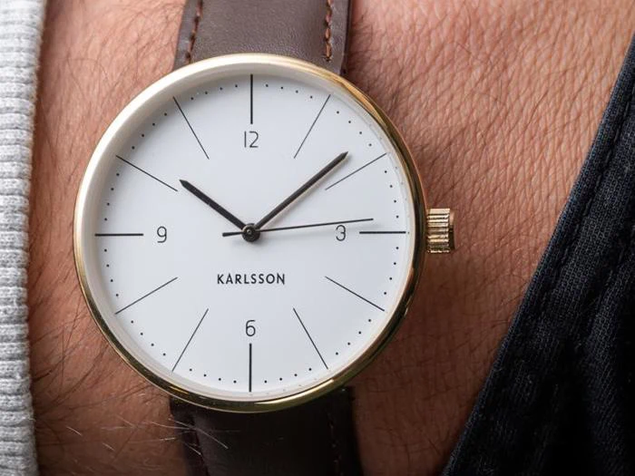 Nærbillede af et håndled iført et analogt Karlsson-ur med guldkant, hvid urskive og brun læderrem, der viser tiden 1:47.