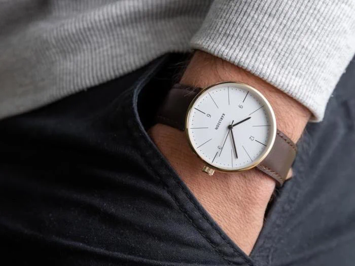 En person iført et minimalistisk armbåndsur med brun læderrem og hvid urskive hviler hånden i lommen på mørke jeans.
