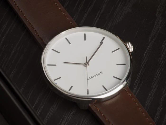Et minimalistisk armbåndsur med hvid urskive, sølvfarvet kasse og brun læderrem, der viser mærket KARLSSON, hviler på en mørk træoverflade.