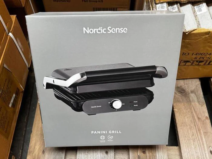 En kasse med en Nordic Sense Panini-grill på en træpalle, omgivet af andre kasser. Billedet af grillen og produktnavnet er trykt på forsiden af emballagen.