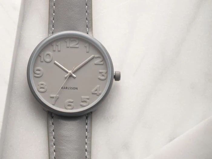 Et gråt Karlsson-armbåndsur med ophøjede tal og grå læderrem hviler på en hvid marmoroverflade.