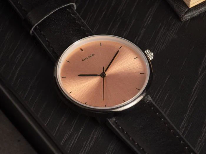 Et minimalistisk armbåndsur med en urskive i rosaguld, sorte timemarkeringer og en sort læderrem, der hviler på en mørk træoverflade.