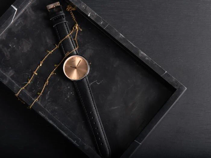 Et guldfarvet armbåndsur med sort læderrem og en fin guldkæde på en sort marmorbakke mod en mørk baggrund.