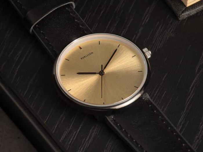 Et minimalistisk armbåndsur med en guldskive, sorte timemarkeringer og en sort læderrem, der hviler på en mørk træoverflade.