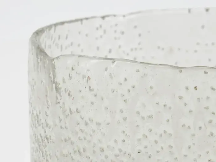 Nærbillede af en klar, struktureret glasbeholder med en bølget kant og hævede prikmønstre på en almindelig hvid baggrund.