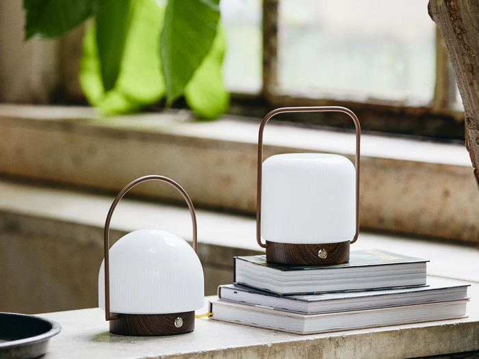 To moderne lanternelignende lamper med træfødder og hvide toppe står i en vindueskarm, den ene oven på stablede bøger, med grønne planter og et vindue i baggrunden.