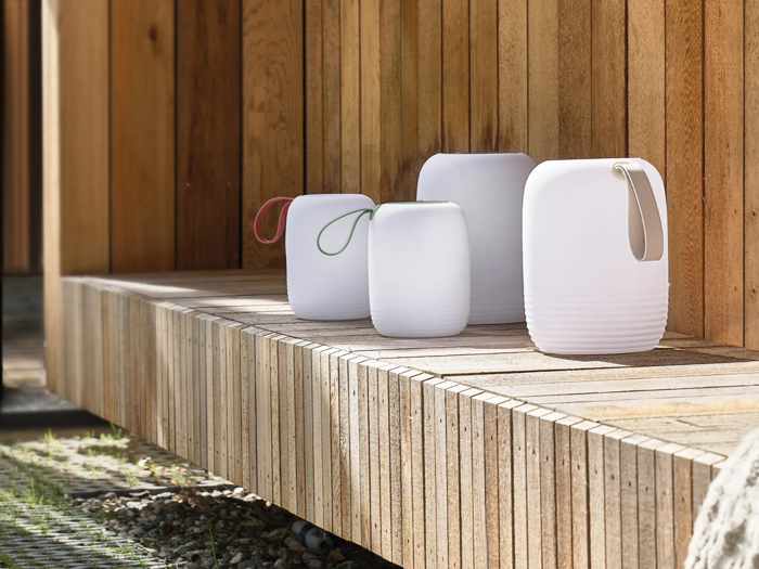 Fire moderne hvide lanterner med farvede håndtag sidder på en træbænk mod en trævæg, hvor sollyset kaster skygger.