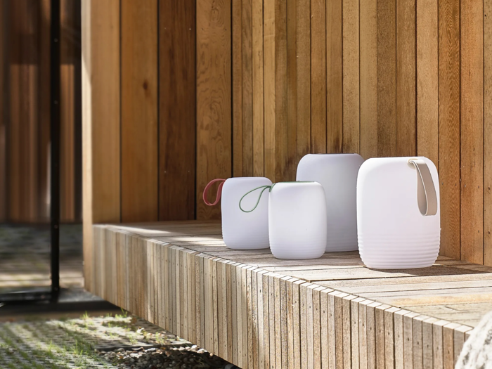 Fire moderne hvide lanterner med farvede håndtag sidder på en træbænk mod en lodret træpladevæg i et udendørs miljø.