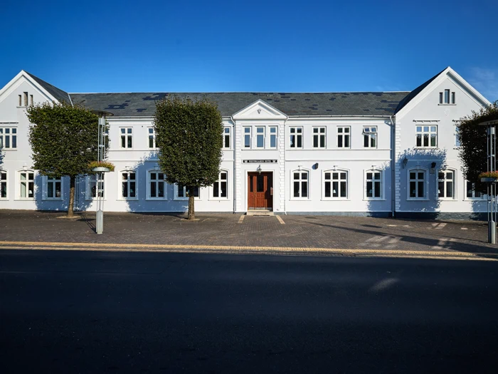 En stor hvid bygning med mange vinduer og en central trædør, flankeret af to grønne træer, under en klar blå himmel.
