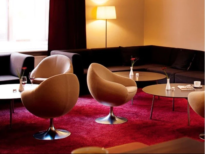 Moderne lounge med beige drejestole, lave borde med blomster, en mørk sektionssofa, rødt tæppe og en tændt gulvlampe i et svagt oplyst rum.