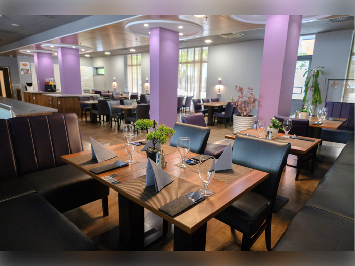 Et moderne restaurantinteriør med lilla vægge, pænt dækkede træborde, foldede servietter, glas og potteplanter. Naturligt lys kommer ind gennem store vinduer i baggrunden.