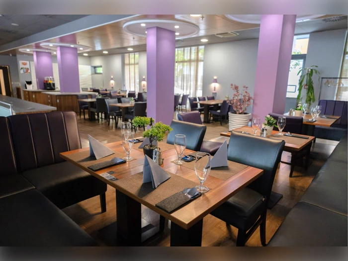 Et moderne restaurantinteriør med lilla vægge, pænt dækkede træborde, foldede servietter, glas og potteplanter. Naturligt lys kommer ind gennem store vinduer i baggrunden.