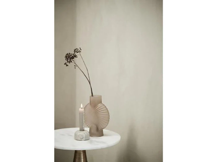 En moderne beige vase med tørrede grene og et tændt stearinlys står på et rundt, hvidt marmorbord mod en ensfarvet, lys væg.