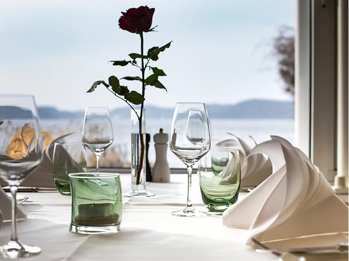 Elegant borddækning med foldede servietter, tomme vinglas, grønne glasskåle og en enkelt rød rose i en vase med en smuk udsigt over søen i baggrunden.