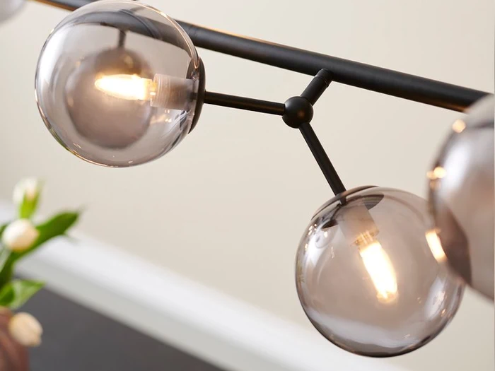 Nærbillede af en moderne sort lampe med runde, røgfarvede glaskugler og synlige pærer, med slørede hvide blomster i baggrunden.